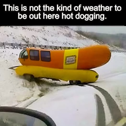 A bright orange and yellow hot dog-shaped vehicle is stuck in snowy terrain, viewed from inside another car. The overlaid…