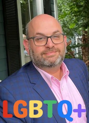 A bald man with glasses and a beard, wearing a blue blazer over a pink shirt, smiles at the camera outdoors. Large, colorful…