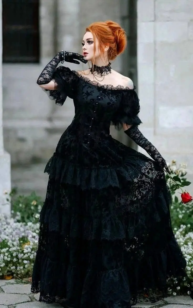 A woman with red hair styled up, wearing a black lace Victorian-style gown with ruffles, long gloves, and a choker, poses…