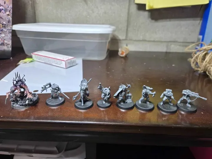 A row of unpainted Warhammer 40k miniatures sits on a wooden desk, with one heavily detailed, colorful model on the far left…