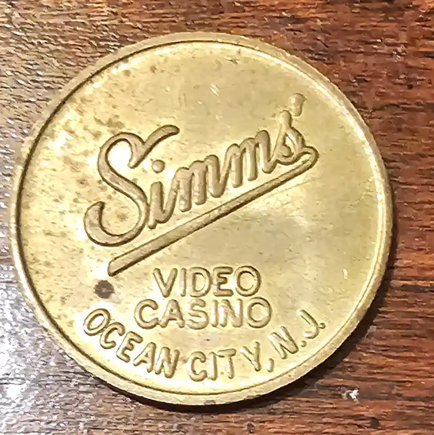 A close-up photo of a gold-colored casino token resting on a wooden surface. The token is embossed with the name 'Simms' in…