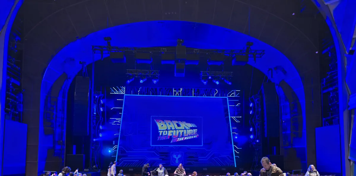 A wide shot of a theater stage bathed in blue light, featuring a large screen displaying the logo for 'Back to the Future…