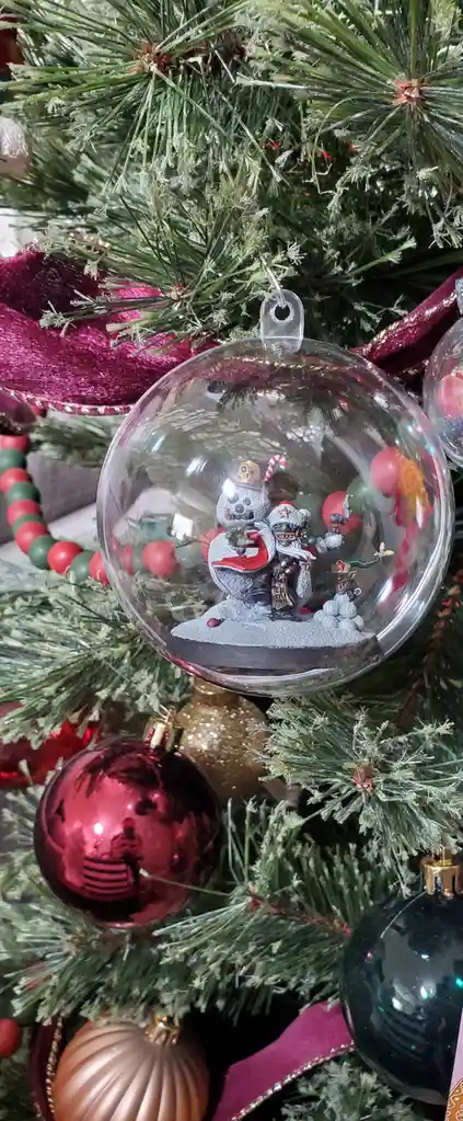 A close-up of a clear Christmas ornament hanging on a decorated tree, containing a miniature diorama of zombie-like figures…