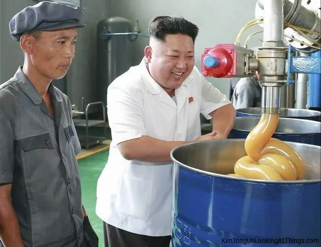 A man in a white shirt smiles broadly while watching thick, yellowish liquid being pumped into a blue barrel, while another…