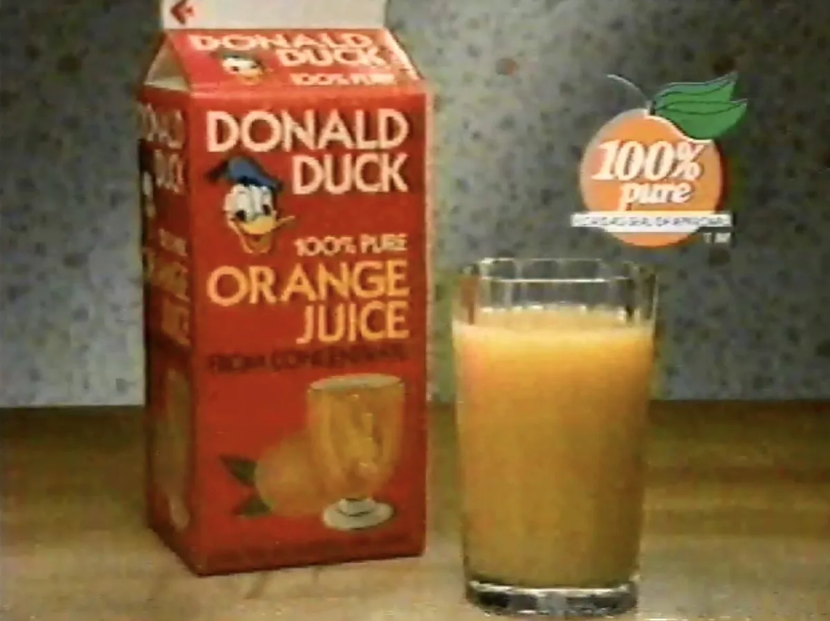 A vintage red carton of Donald Duck brand orange juice stands next to a glass filled with the juice. The carton features…