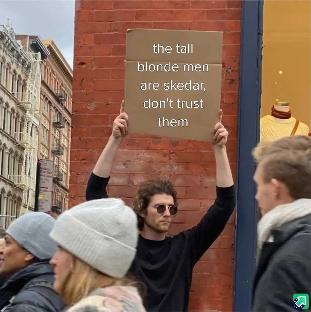 A man with dark hair and sunglasses holds up a cardboard sign in front of a brick wall, displaying the text 'the tall blonde…