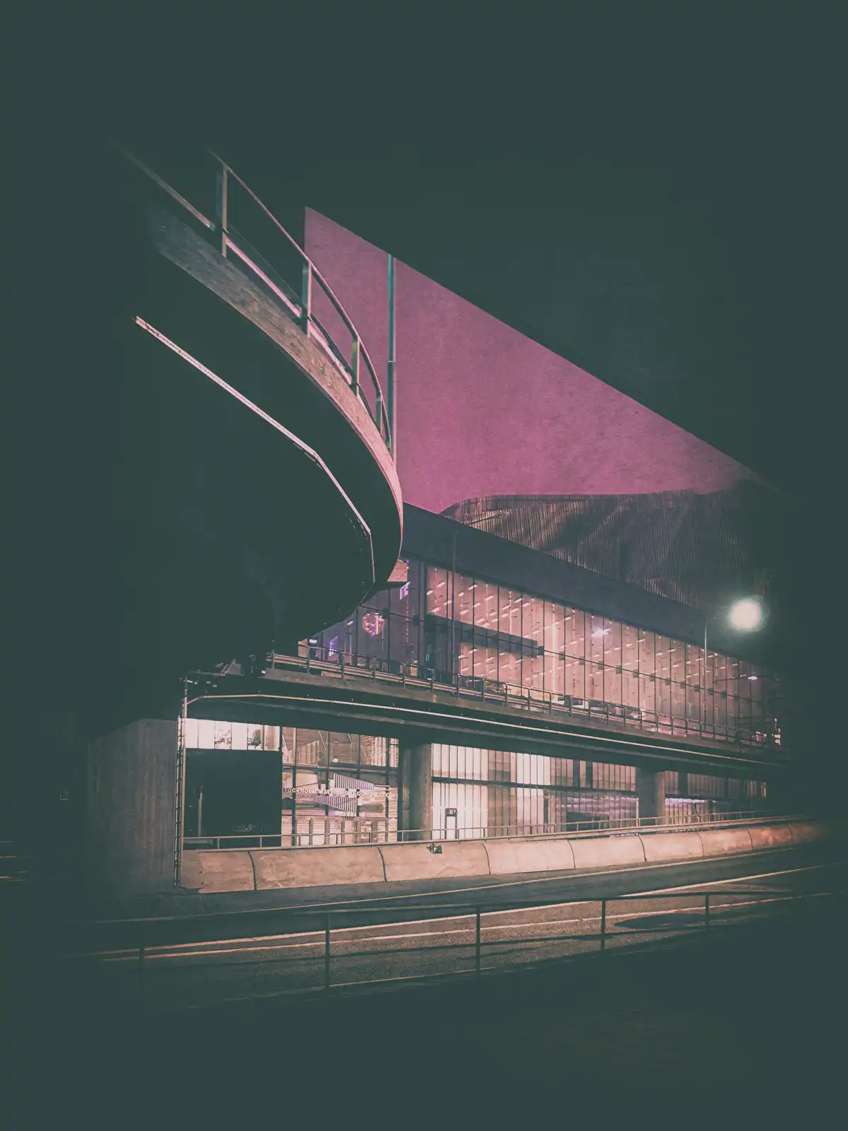 A low-angle night photograph of a modern, angular building with a curved elevated walkway, illuminated by artificial lights…