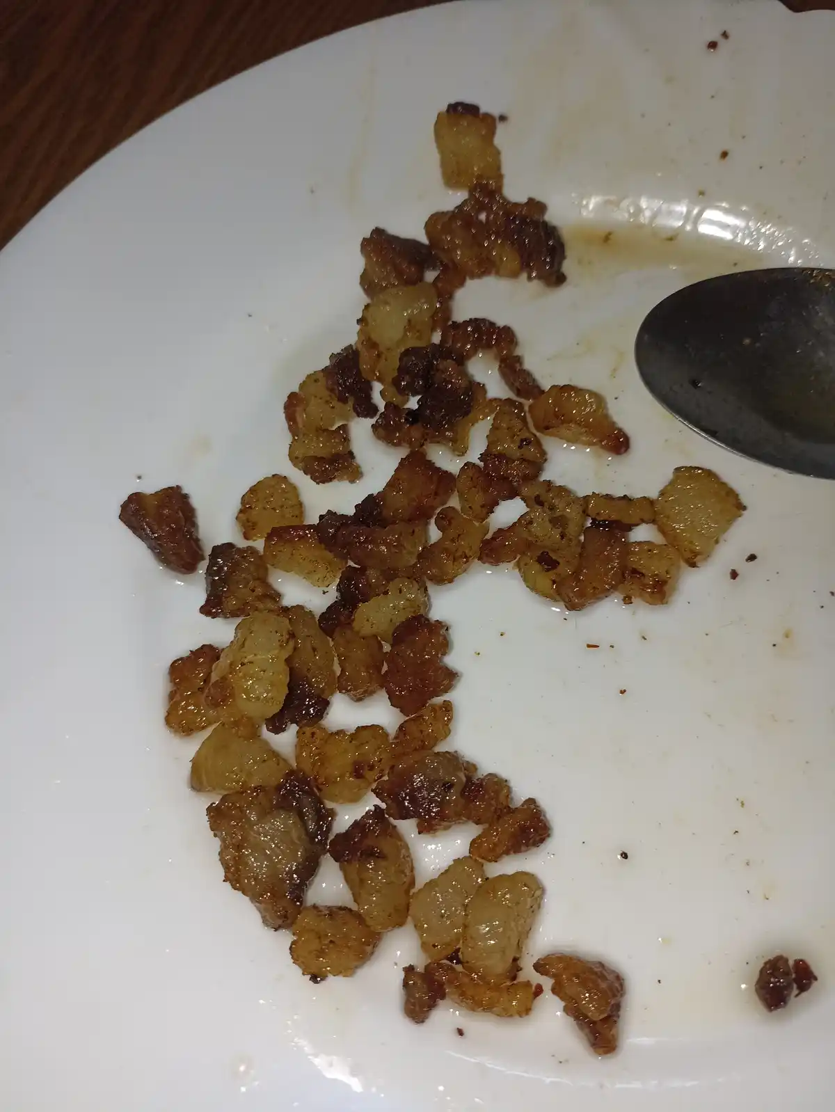 A close-up photo shows a white plate with scattered, crispy, golden-brown bacon bits and grease, with a metal spoon resting…