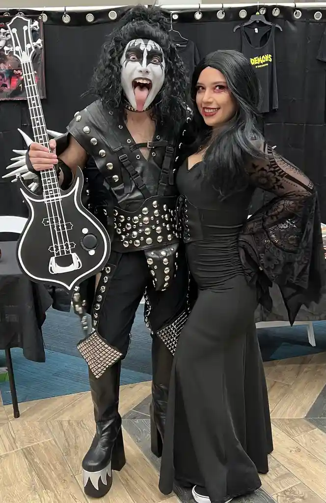 Two people dressed in elaborate rock costumes pose for a photo. The person on the left wears a full-face white and black…