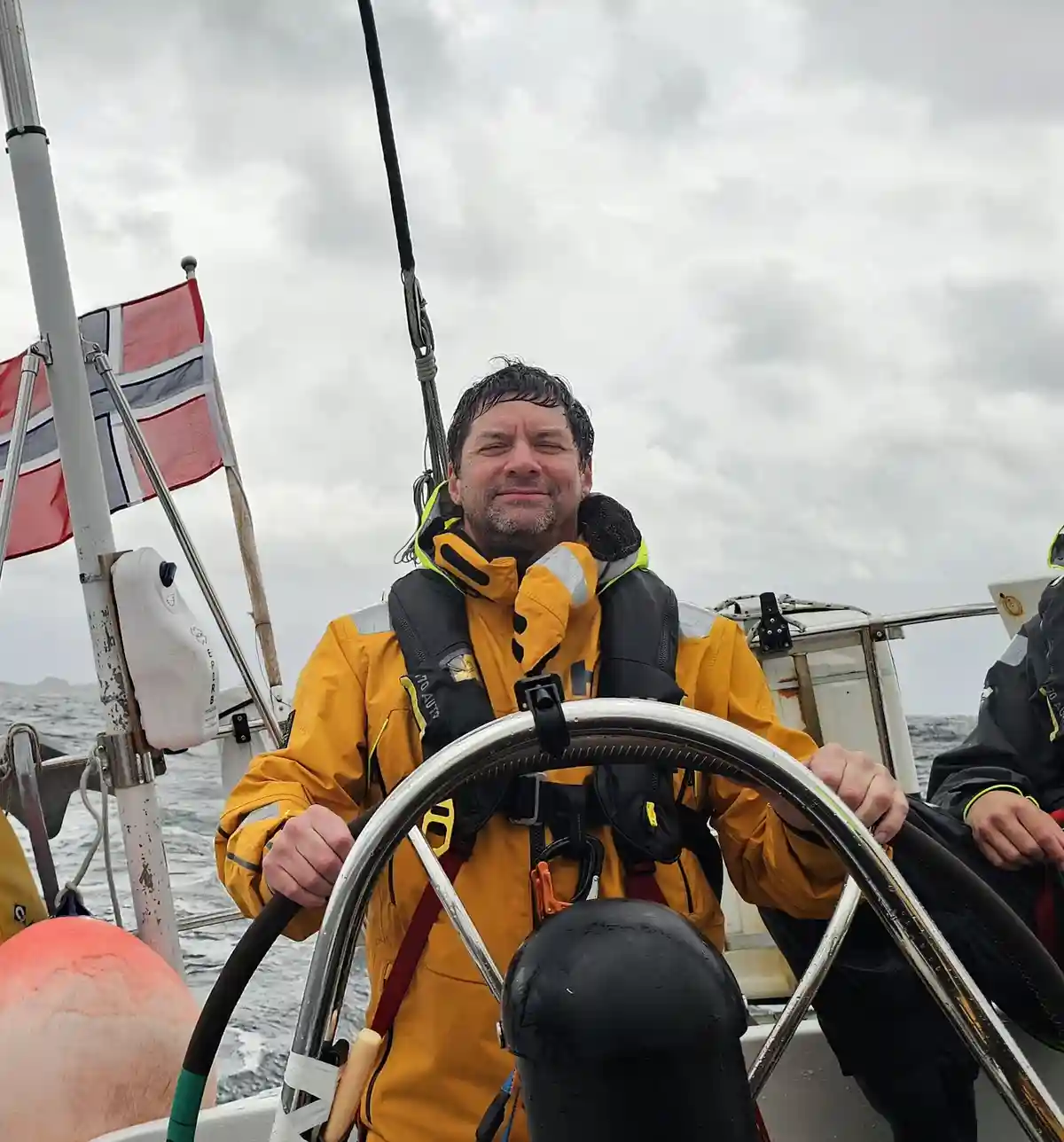 A man in a yellow waterproof jacket and life vest is steering a boat, looking tired with his eyes half-closed. Behind him, a…