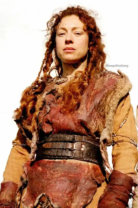 A woman with curly red hair and braids wears a tattered, fur-trimmed outfit that appears to be made of raw meat, with a…