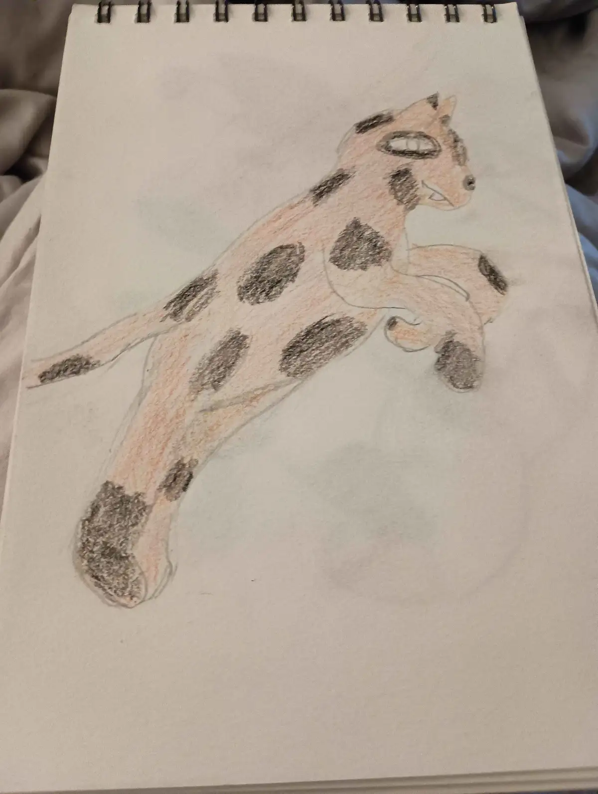 A child's colored pencil drawing on a spiral-bound notebook shows a cat with large black spots covering its body, resembling…