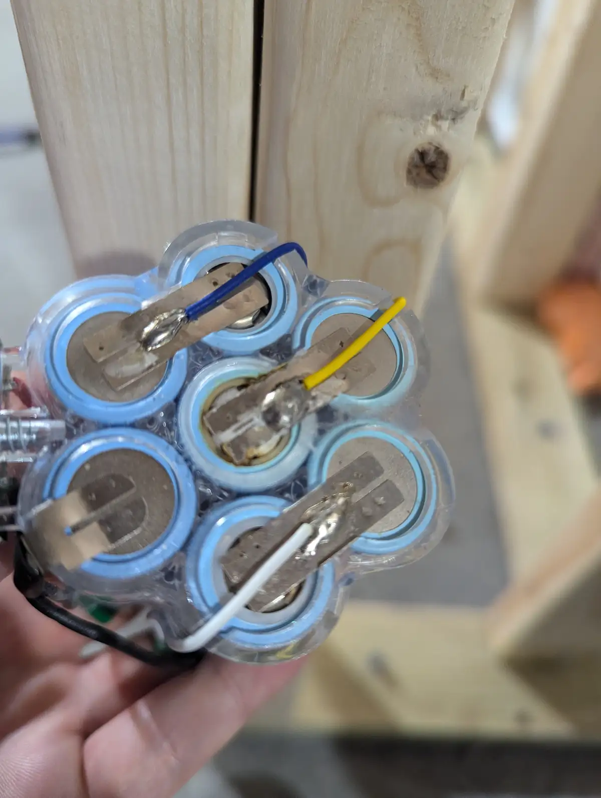 A hand holds a clear plastic battery pack containing six cylindrical batteries, some with visible soldered wires (blue…