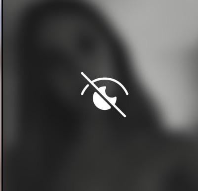 A grayscale, heavily blurred image of a person's face, with a white 'no eye' symbol overlaid in the center. The symbol is a…