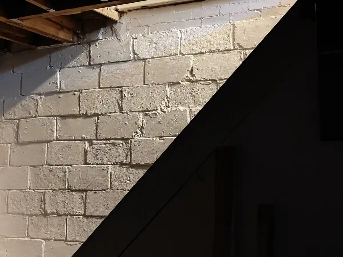 A dimly lit photo shows a white-painted brick wall with exposed wooden beams above and a dark diagonal beam cutting across…