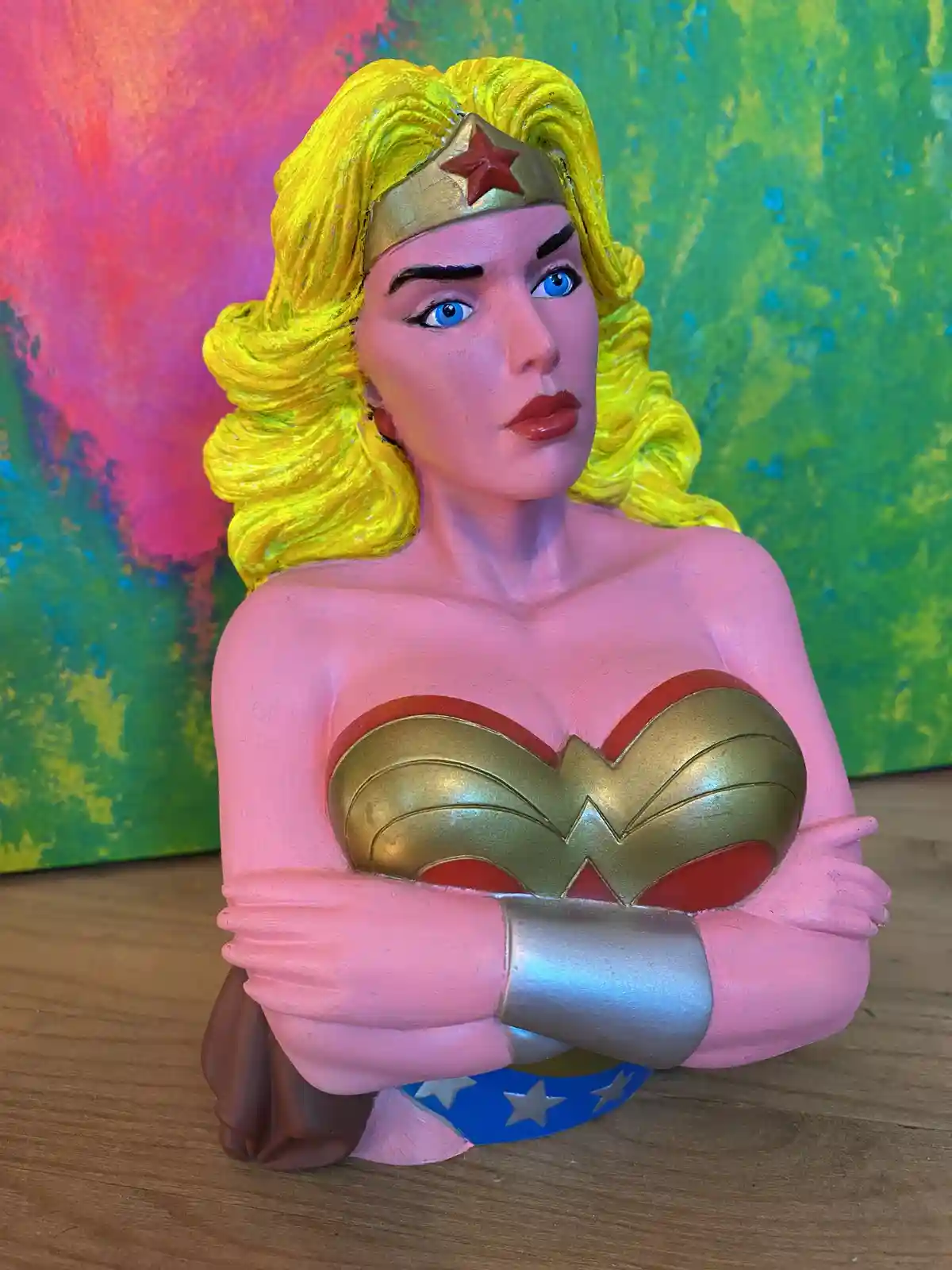 A painted bust of Wonder Woman with her arms crossed, standing on a wooden surface in front of a colorful abstract…