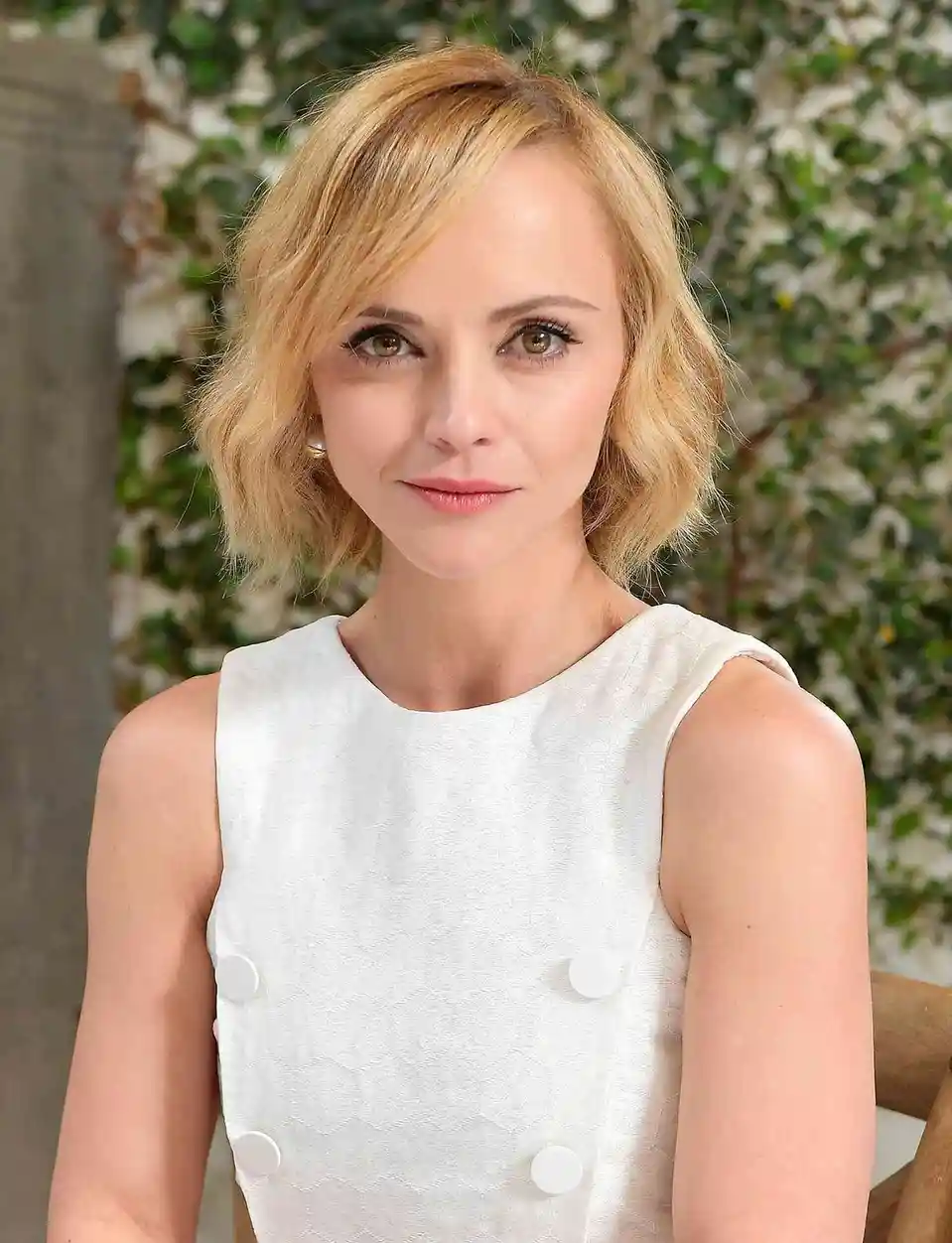 A woman with short blonde hair and a white sleeveless dress with large buttons looks directly at the camera with a subtle…