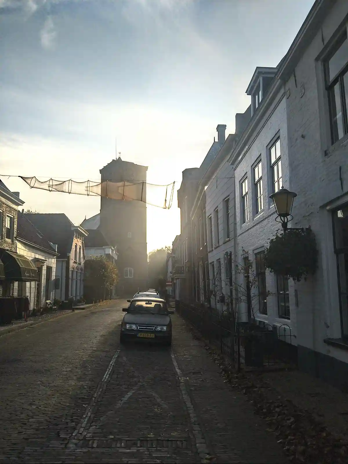 A cobblestone street in a European town with historic buildings, a church tower in the distance, and a car parked in the…