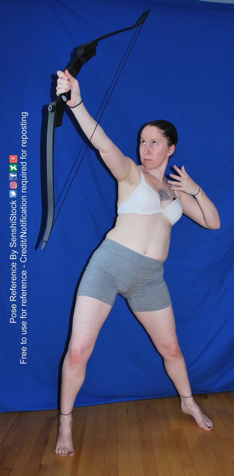 A woman in a white sports bra and grey shorts stands barefoot on a wooden floor against a blue backdrop, holding a black…