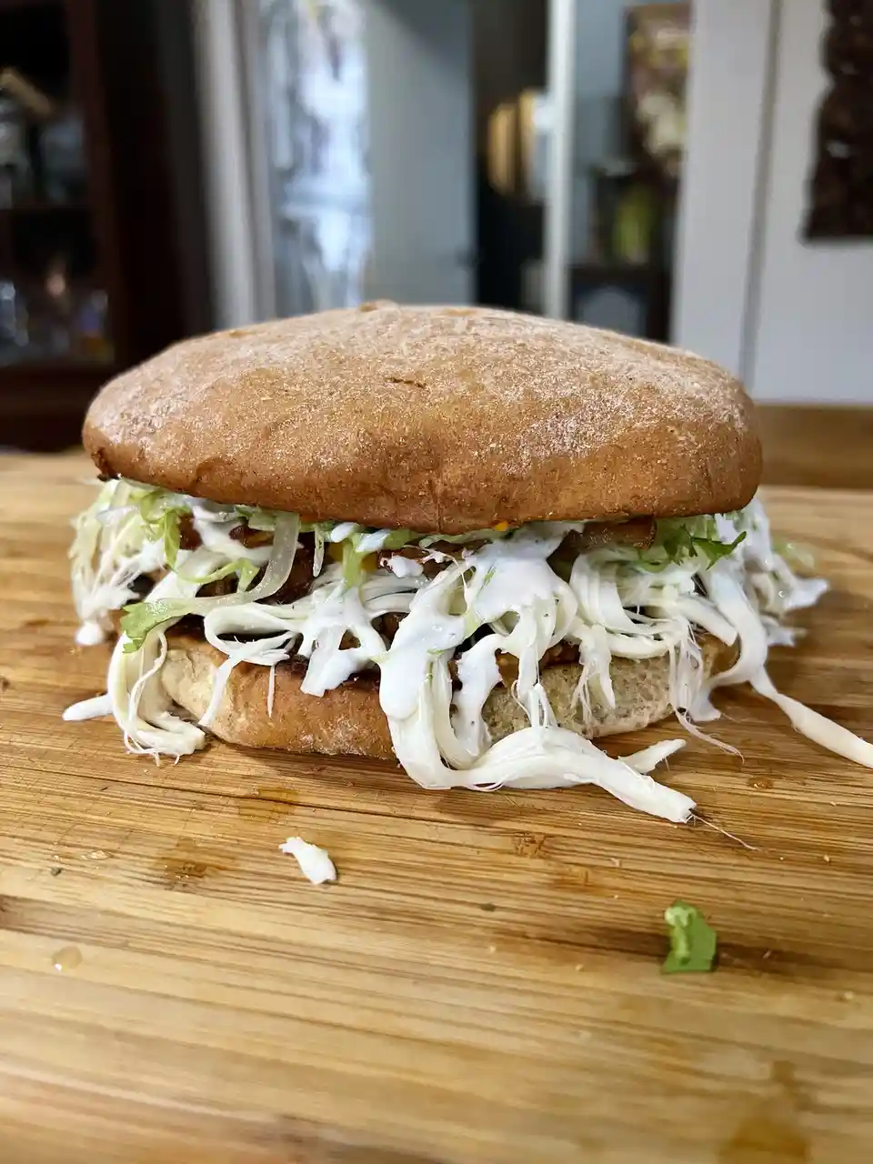 A close-up photo shows a large, messy sandwich on a wooden cutting board, overflowing with shredded lettuce, a creamy white…
