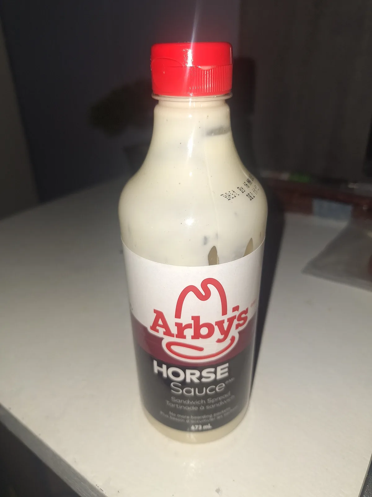 A close-up photo shows a plastic bottle of Arby's sauce with a red cap and white label, sitting on a white countertop. The…