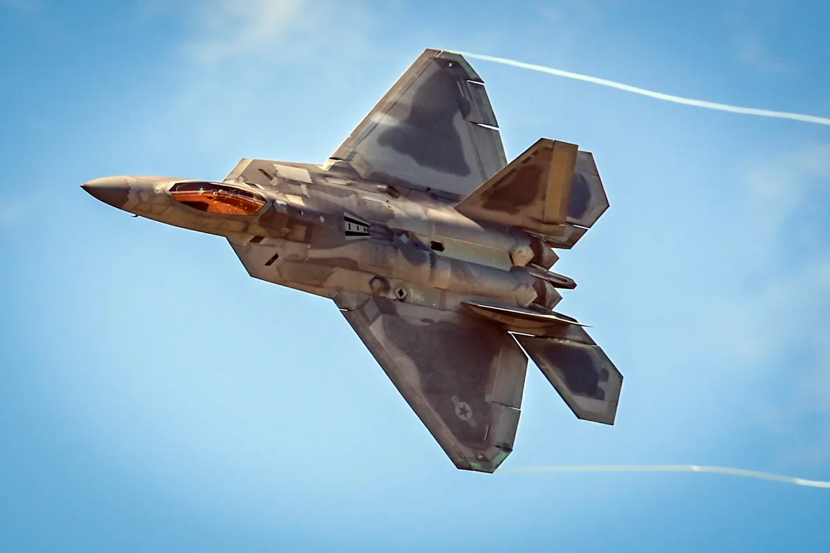 A fighter jet, likely an F-22 Raptor, is captured in flight against a clear blue sky, banking sharply with visible…
