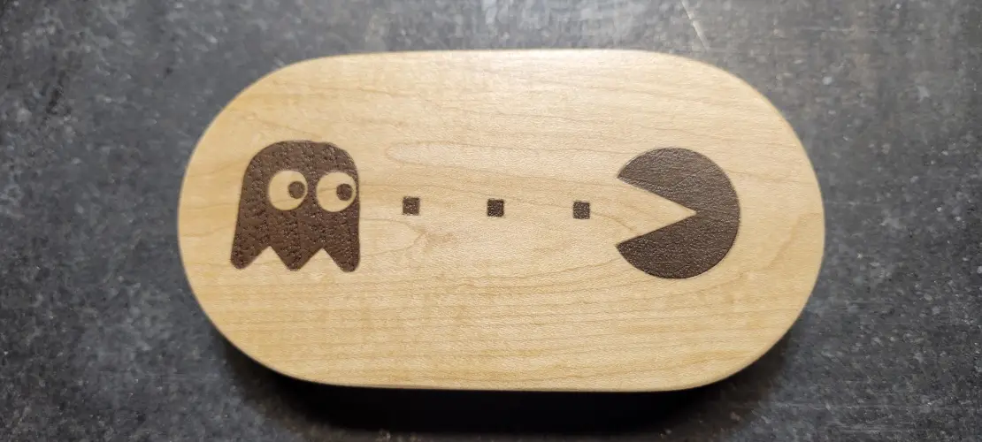 A light-colored wooden oval object features a laser-etched Pac-Man character on the right and a ghost on the left, with…