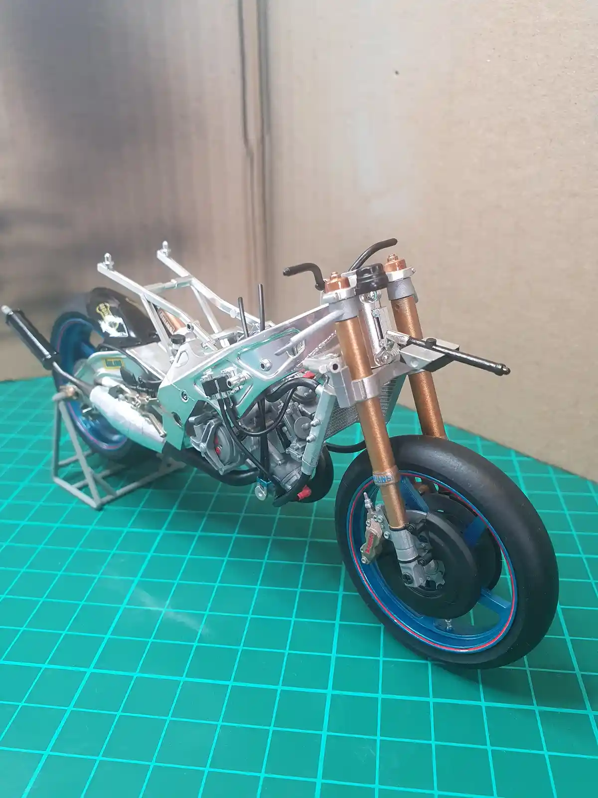 A highly detailed scale model of a motorcycle, mostly assembled but missing its body panels, sits on a green grid mat. The…