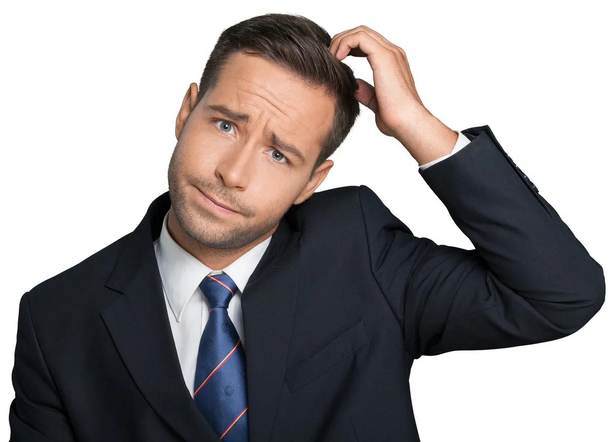 a man in a dark suit and blue tie with a confused expression, scratching his head with his right hand against a white…