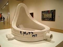 A white ceramic urinal is displayed on a pedestal in a museum gallery, with the text 'R. MUTT 1917' handwritten in black…