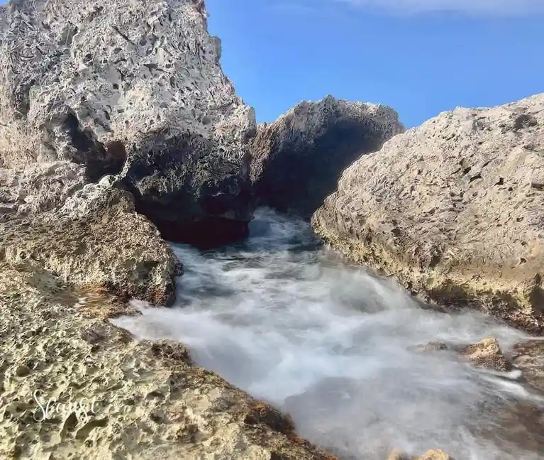 A long-exposure photograph shows turbulent ocean water flowing through a narrow, rocky cove under a clear blue sky. The…