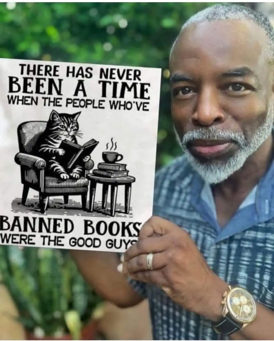 A man with a gray beard and plaid shirt holds up a sign featuring a black-and-white illustration of a cat reading a book in…
