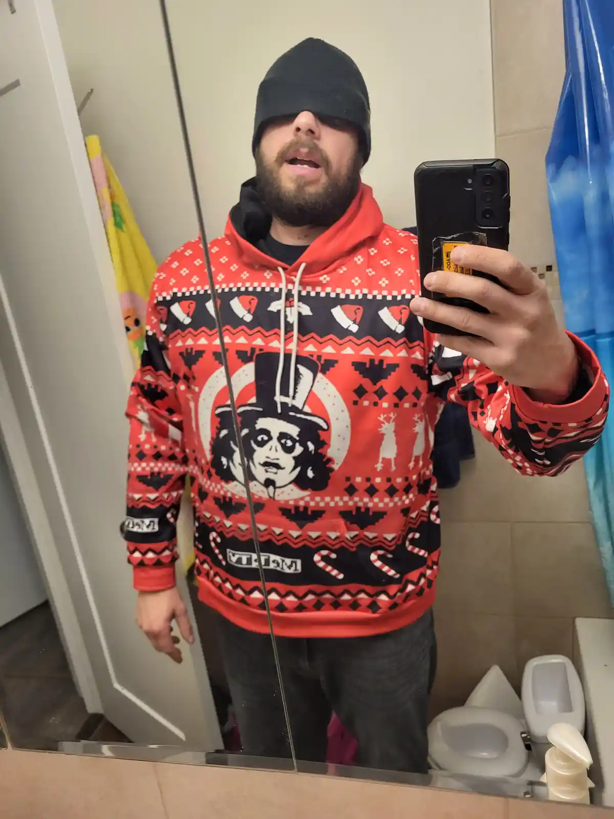 A man with a beard and black beanie takes a mirror selfie in a bathroom while wearing a red and black holiday-themed hoodie…