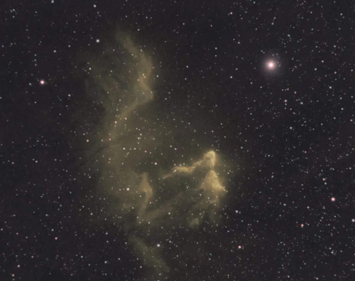 a deep space photograph shows a dark sky filled with countless stars and a prominent yellowish nebula. The nebula's wispy…