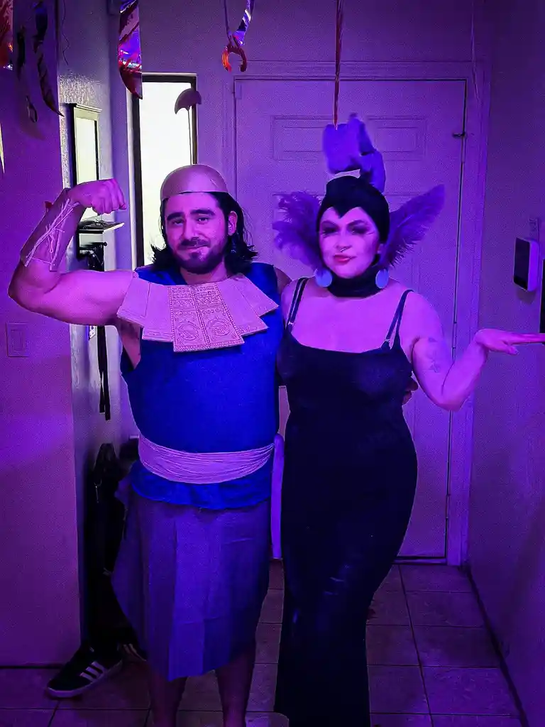 Two people in costumes stand in a purple-lit hallway. The person on the left, wearing a blue Egyptian-style outfit with a…