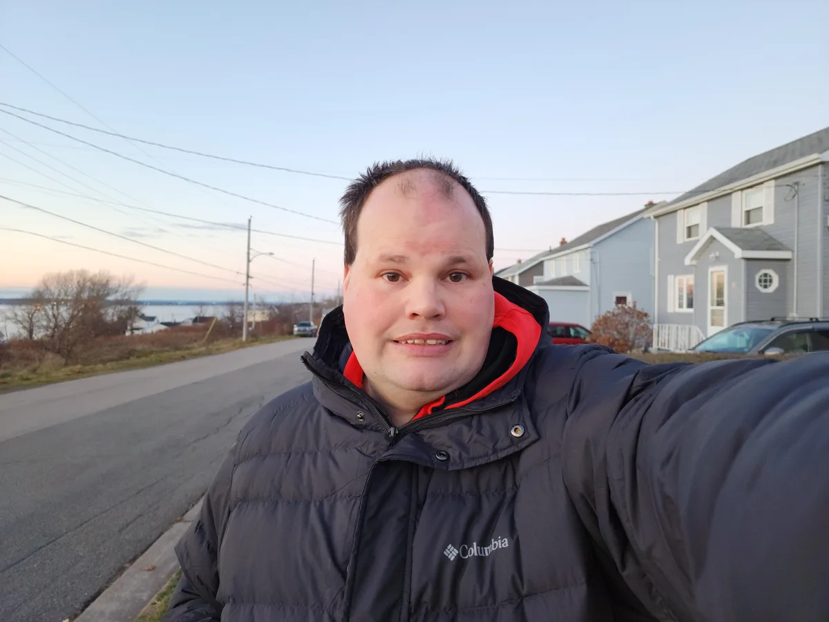 A man in a dark Columbia puffer jacket with a red-lined hood takes a selfie outdoors on a residential street at dusk. He has…
