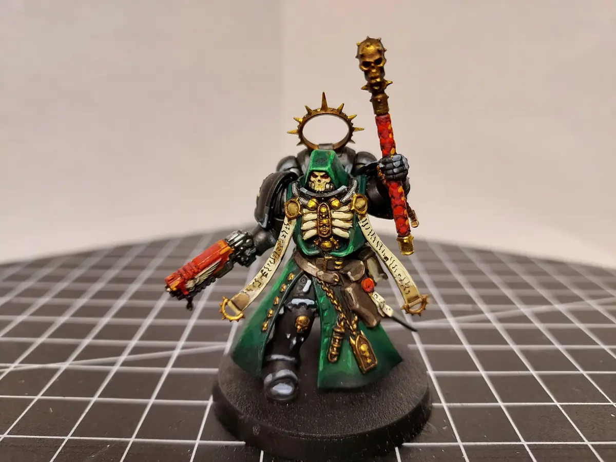 A painted miniature of a green-robed, skull-faced warrior in dark armor stands on a black circular base, holding a red and…