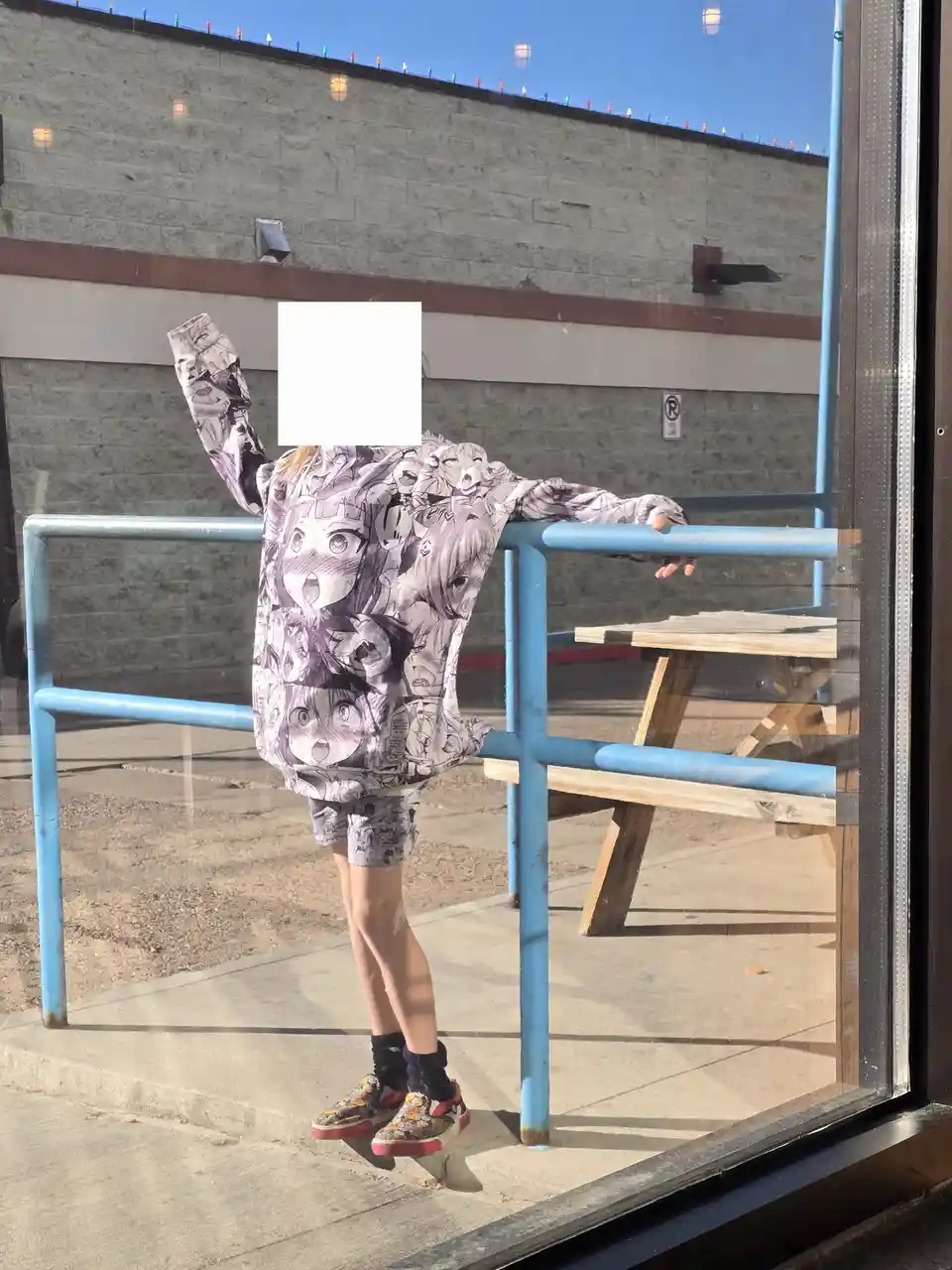 A person is seen through a glass window, posing with one arm raised and the other resting on a blue railing. They are…