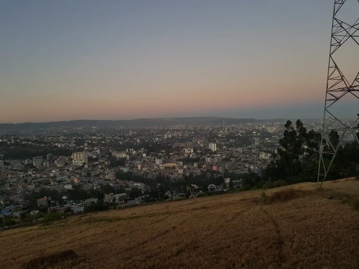 A scenic photo taken from a hillside at dusk, showing a sprawling cityscape below with buildings and distant hills under a…