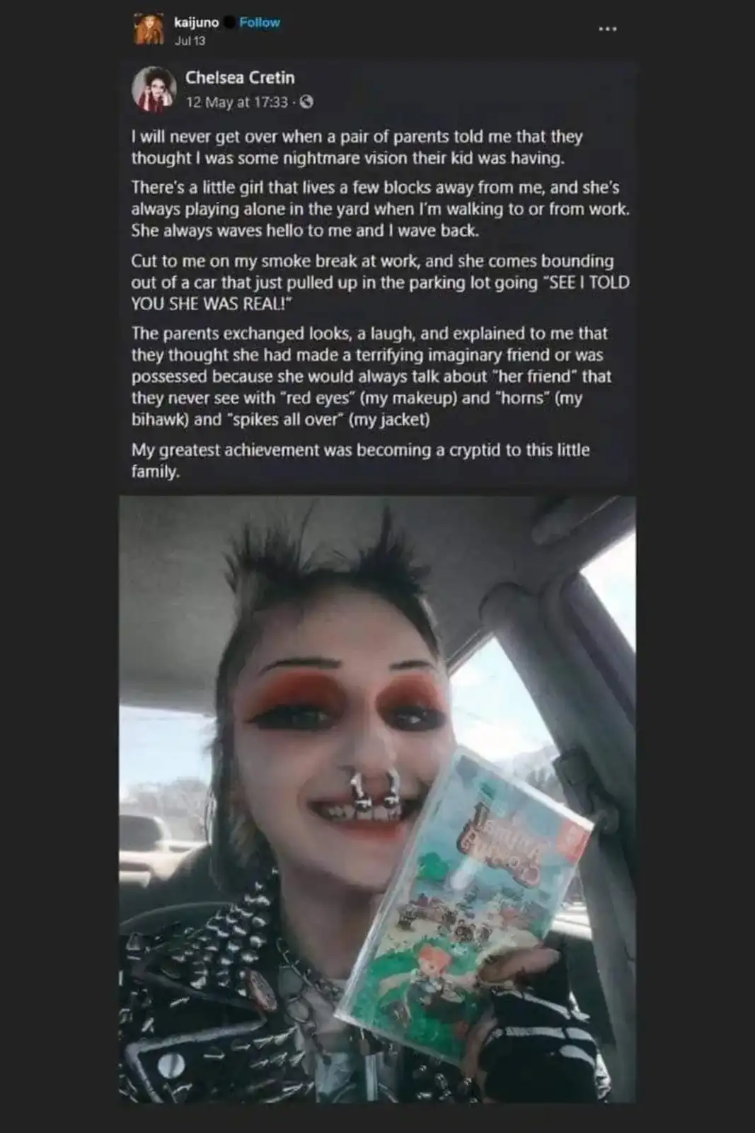 A person with spiky hair, red eye makeup, a nose ring, and a studded jacket is smiling while holding a 'Tamagotchi' game…