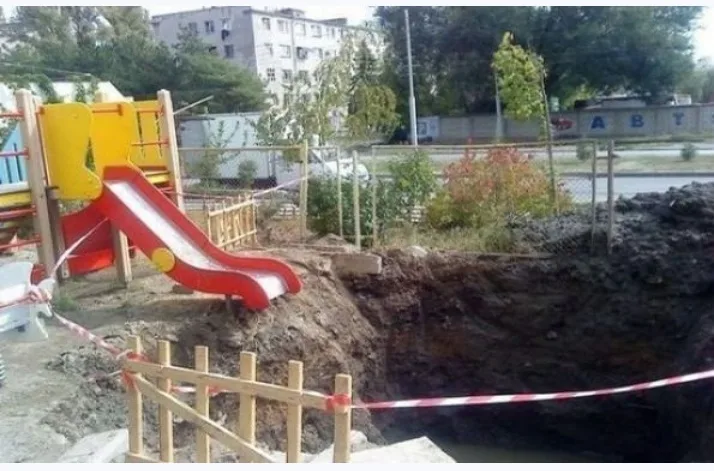 a red and yellow playground slide is perched precariously on the edge of a large, freshly dug hole, cordoned off with red…