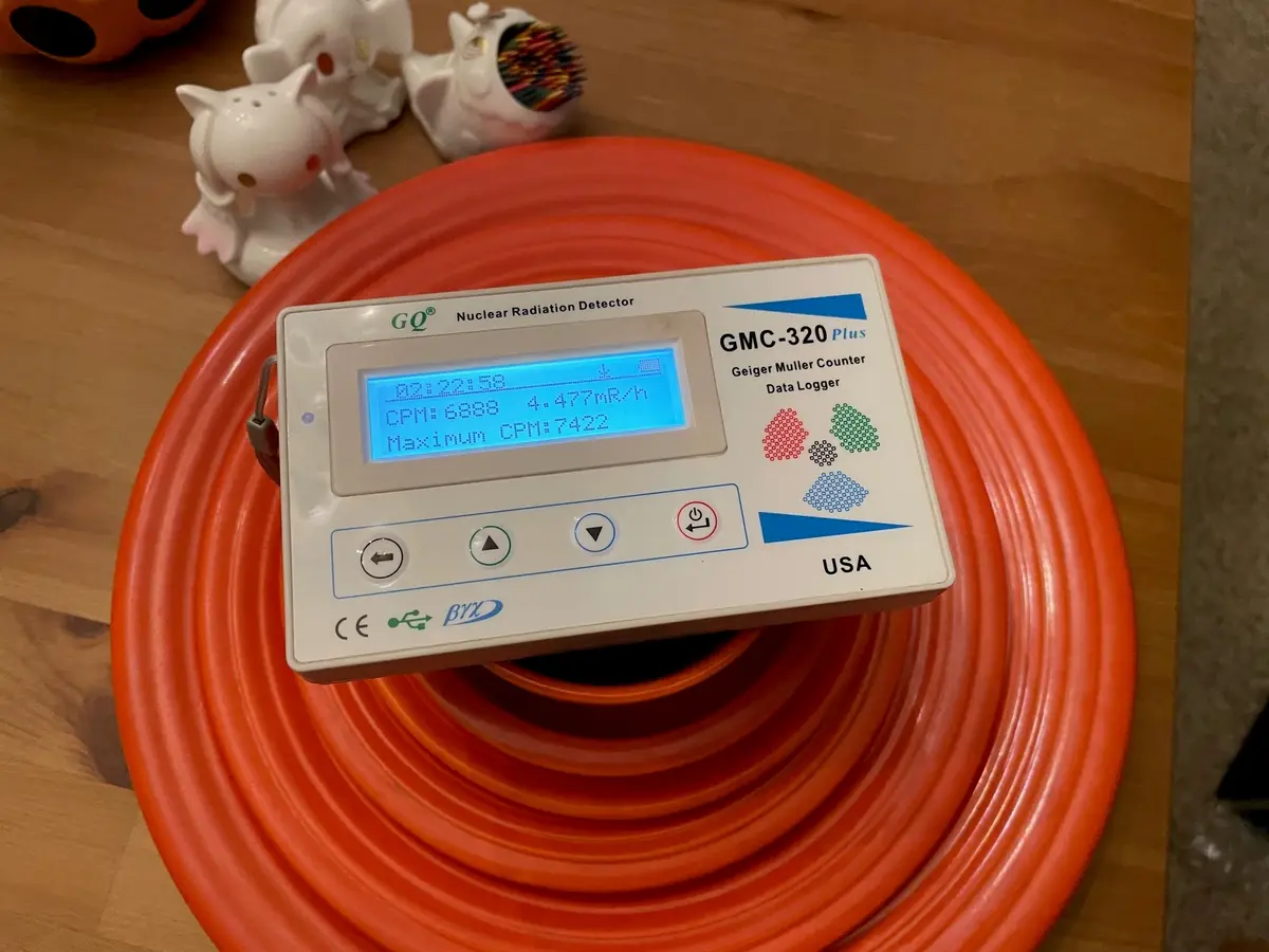 A white GMC-320 Plus Geiger Muller Counter sits atop a coiled orange hose on a wooden surface, its blue LCD screen…