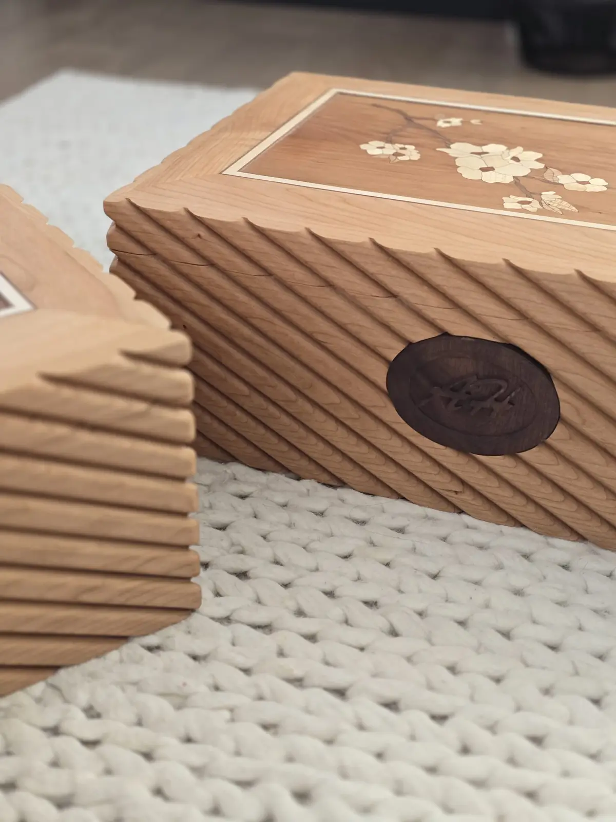 A close-up photo shows two light-colored wooden boxes with decorative inlaid cherry blossom patterns on top and grooved…