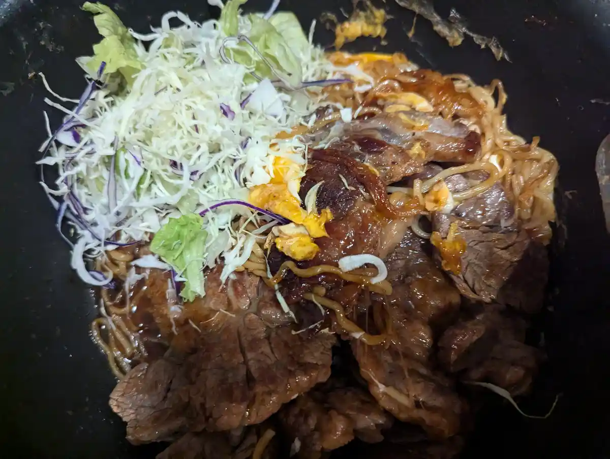 a close-up photo of a savory dish in a black pan, featuring cooked beef slices, stir-fried noodles, and a pile of shredded…
