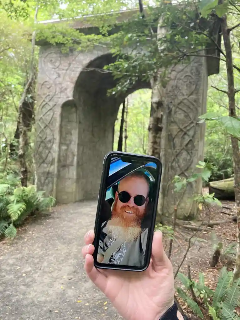 A hand holds a smartphone in front of a stone archway in a forest, displaying a selfie of a smiling man with a beard and…