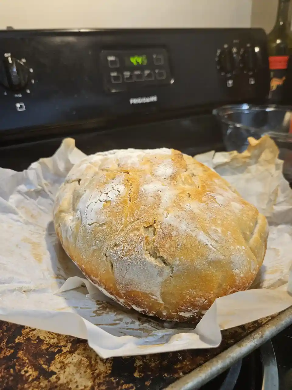 A freshly baked, golden-brown round loaf of bread sits on parchment paper atop a metal baking sheet, placed in front of a…