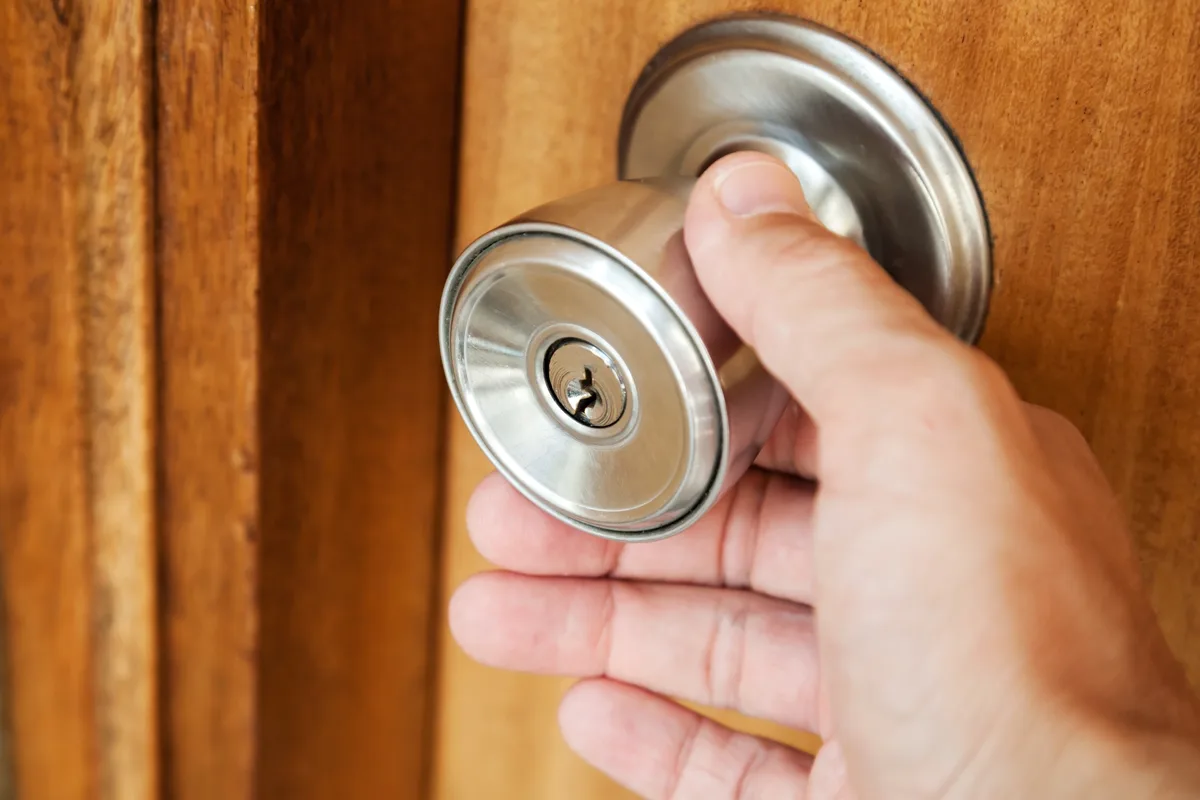 A close-up photo shows a person's hand gripping a shiny, metallic doorknob on a wooden door. The doorknob has a visible…