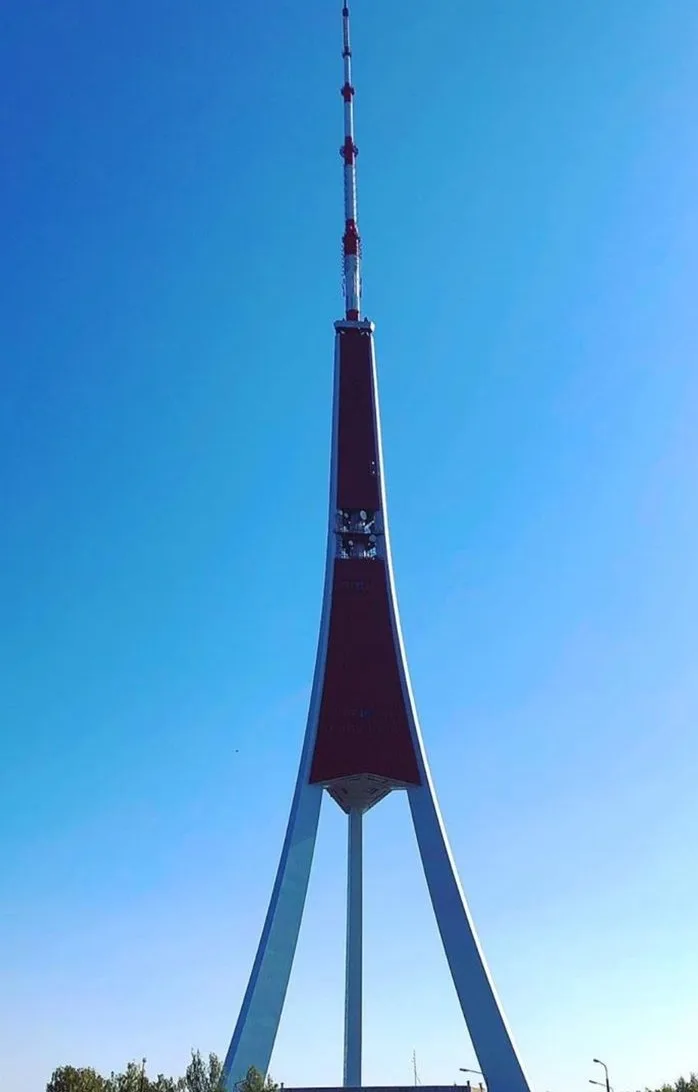 A tall, slender tower with a red and white design and three blue support legs stands against a clear blue sky. The overlaid…