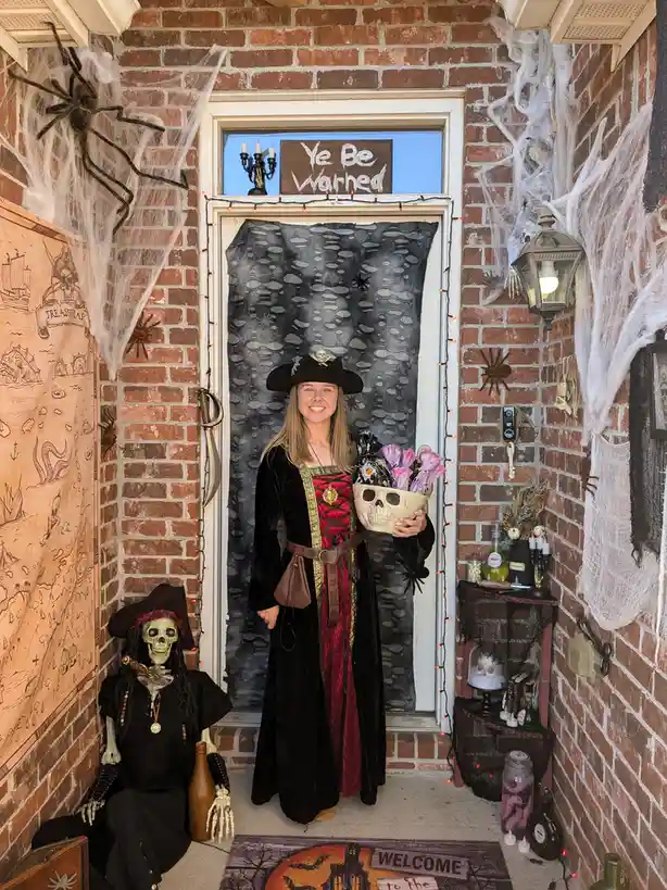 A smiling woman in a pirate costume stands in a heavily decorated Halloween doorway, holding a skull-shaped basket of candy…