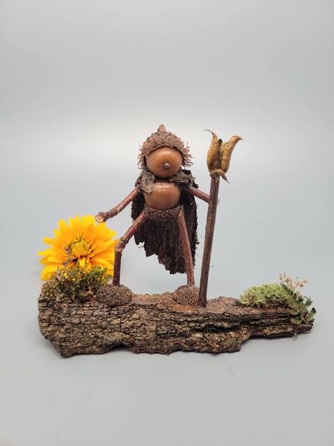 A whimsical handmade figurine crafted from natural materials like acorns, twigs, and moss, standing on a piece of bark with…